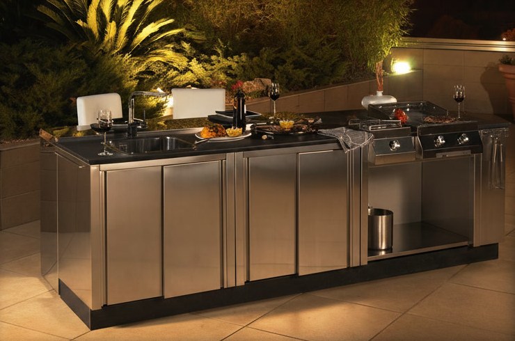 Cucina da esterno tutta in acciaio inossidabile con isola, piano scuro, lavello e piastre, illuminata di sera