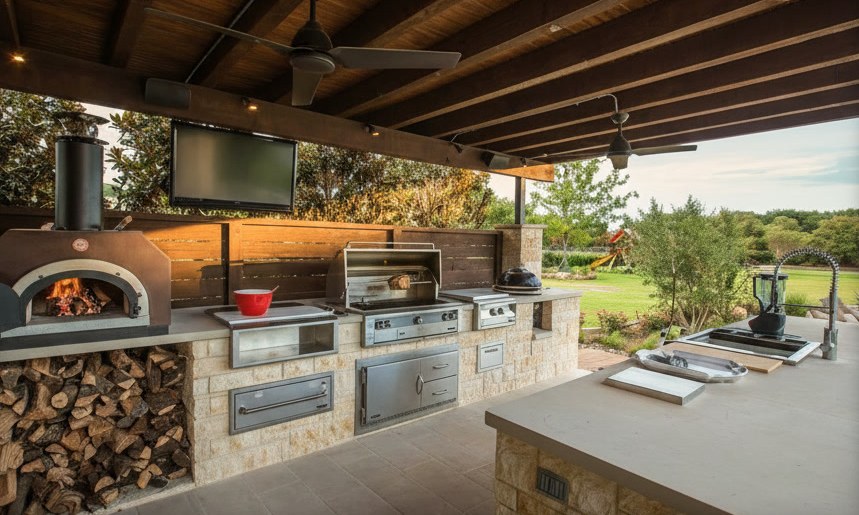 Cucina da esterno in pietra chiara sotto una tettoia in legno, con forno a legna acceso, legnaia, barbecue e isola