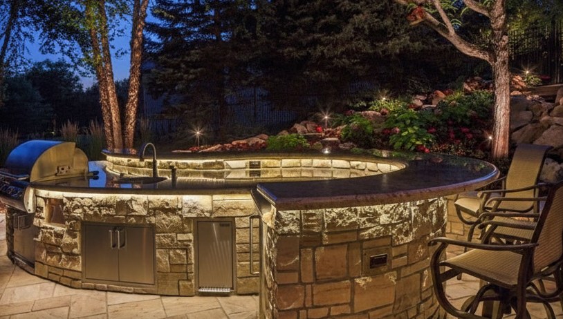 Cucina da esterno circolare in marmo e granito con base rivestita di pietra, barbecue in acciaio e luci accese di sera