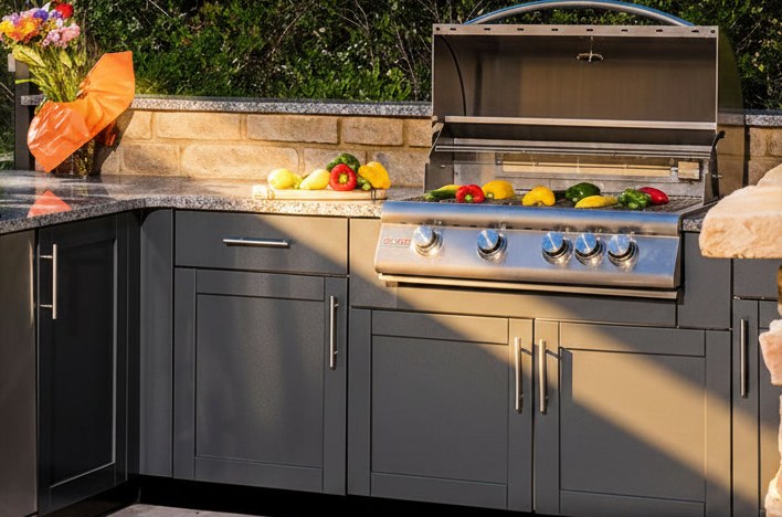 Cucina da esterno ad angolo con piano in granito scuro, barbecue a gas in acciaio incassato e ante grigie