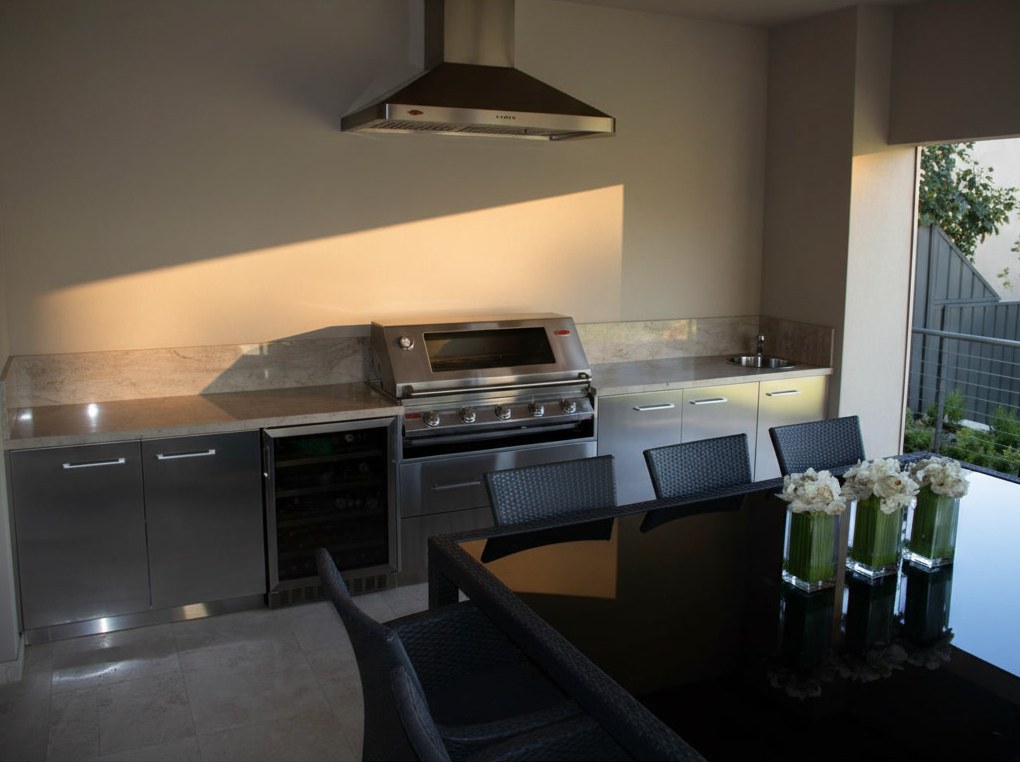 Kitchen under a porch with steel base units, pale marble worktop, wine cooler, gas barbecue and steel hood