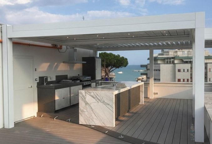 Terrazza con isola chiara venata a cascata e cucina a parete sotto una pergola bianca a lame, con il mare sullo sfondo