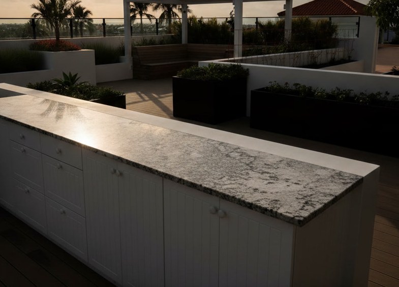 Cucina da esterno con piano in granito su basi bianche, terrazza con fioriere, palme e pergola al tramonto