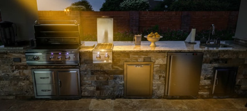 Cucina da esterno lineare rivestita in pietra con piano chiaro, barbecue, fornello, lavello e sportelli in acciaio, di sera