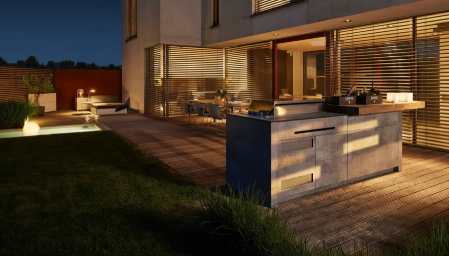Isola cucina da esterno effetto pietra con barbecue su terrazza in legno, casa moderna illuminata al crepuscolo