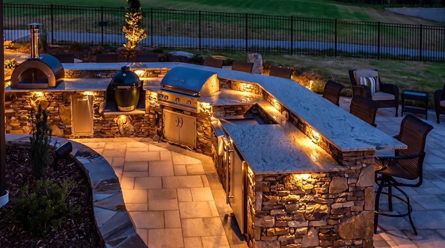 Visione di progetto: grande cucina da esterno curva in pietra con piani in granito illuminati, forno per pizza e barbecue, di notte