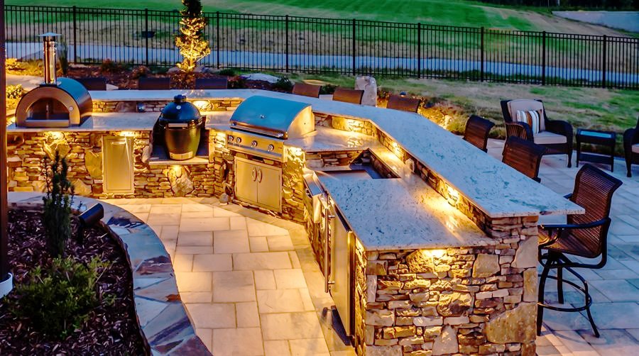 Visione di progetto: grande cucina da esterno curva in pietra con piani in granito illuminati, forno per pizza e barbecue, di notte