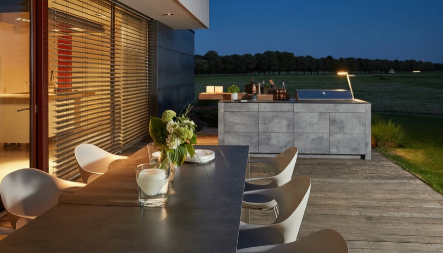 Tavolo da pranzo su terrazza al tramonto con cucina da esterno effetto pietra e paesaggio verde sullo sfondo