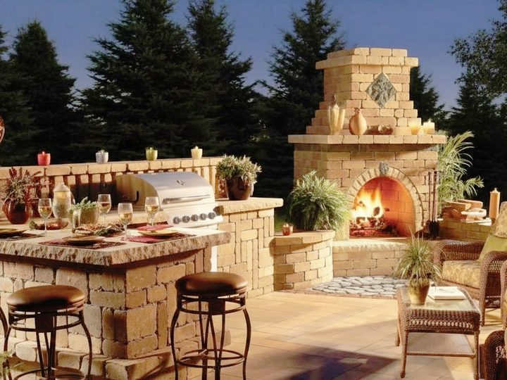 Outdoor kitchen in rough stone with professional barbecue and grand stone fireplace, counter with stools, at dusk