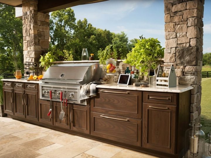Outdoor kitchen in granite and walnut wood with hooded gas barbecue, between stone columns and lawn