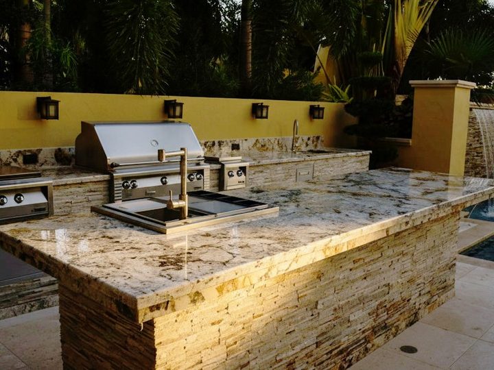 Outdoor kitchen with granite worktop on a stacked-stone base, gas barbecue, sink and a pool waterfall behind