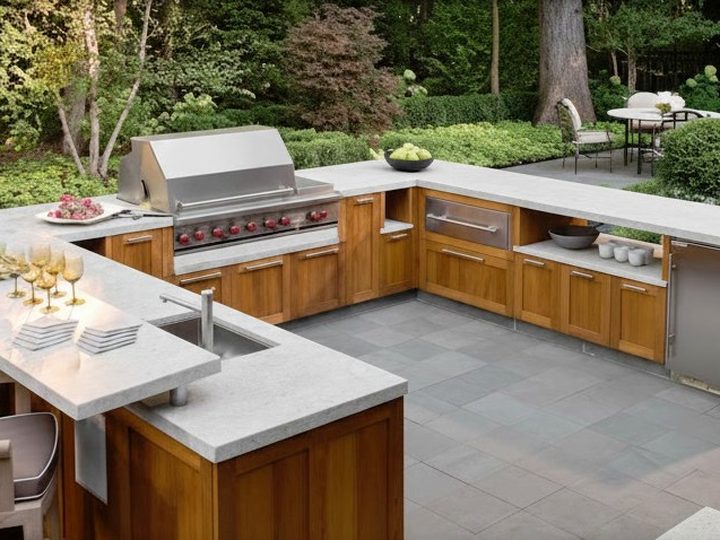 Cucina da esterno con elettrodomestici da incasso, pianale in granito chiaro e struttura in legno trattato, a U
