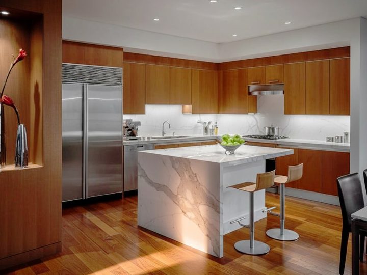 Warm lacquered wooden kitchen with central veined marble table and stainless steel fridge