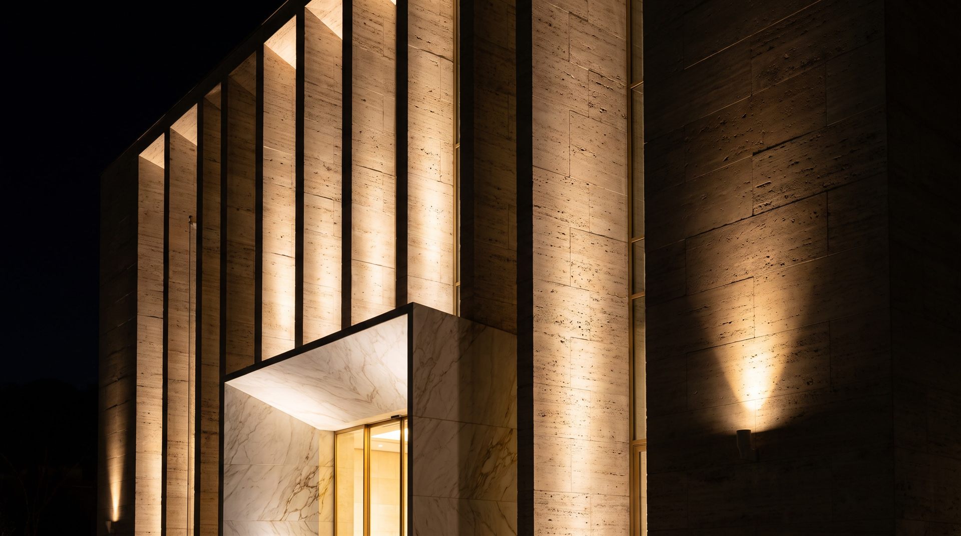 Visione di progetto: facciata monumentale a lesene verticali di travertino romano illuminata dal basso, con portale d'ingresso in marmo