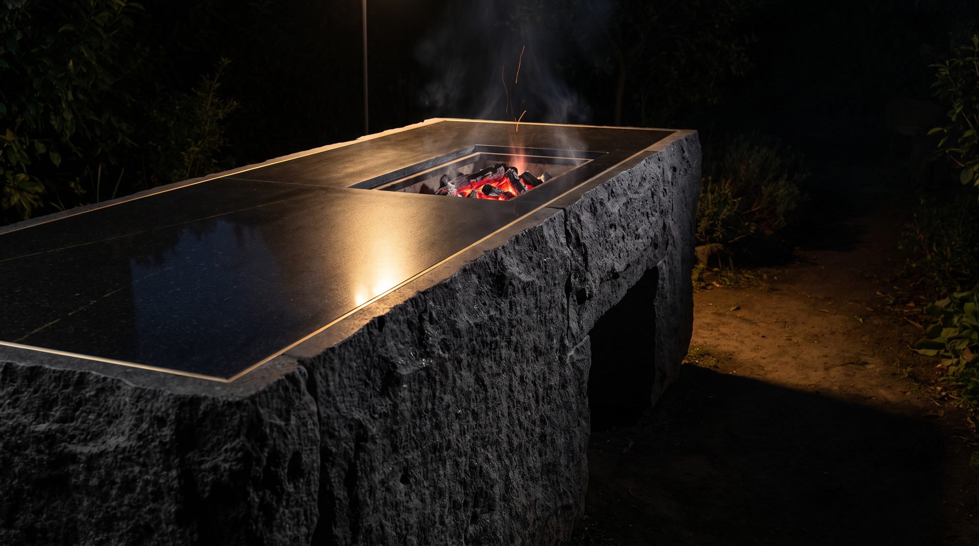 Visione Michelessi: isola in pietra lavica a spacco con piano in granito nero lucido e braciere a carbone vivo incassato, di notte in giardino