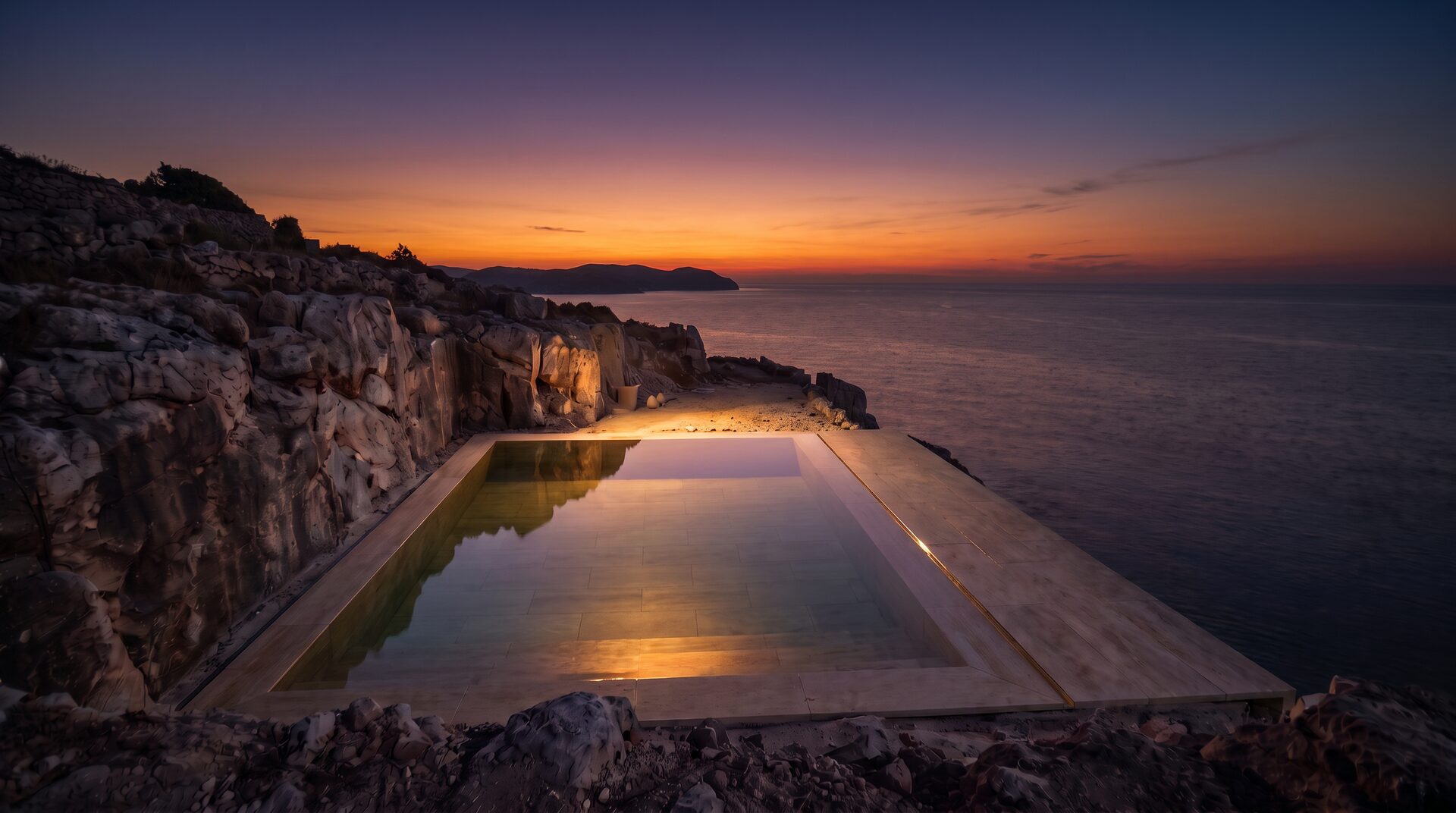 La piscina piena al tramonto: l’acqua a filo del bordo specchia il cielo d’ambra sopra il mare
