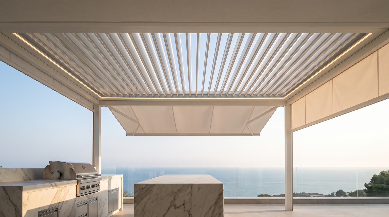 Vista dal basso di una pergola bioclimatica bianca con le lame aperte al sole, luci a led integrate e tenda a bracci; sotto, l'angolo di un'isola da esterno in marmo Calacatta con griglia in acciaio