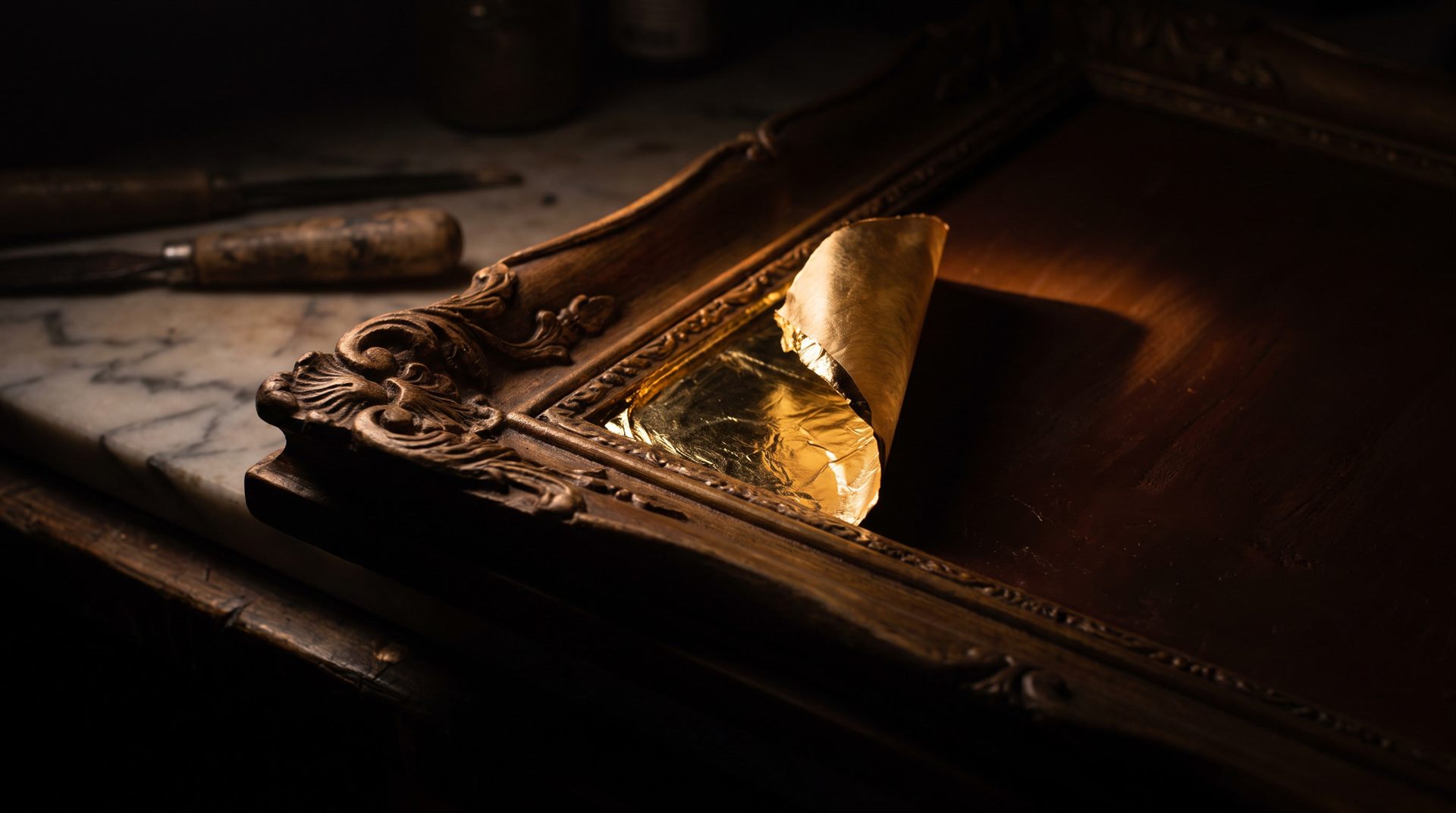 Visione di progetto: primo piano di una foglia d'oro che si solleva dall'angolo intagliato di una cornice, sul banco di marmo del laboratorio nel buio
