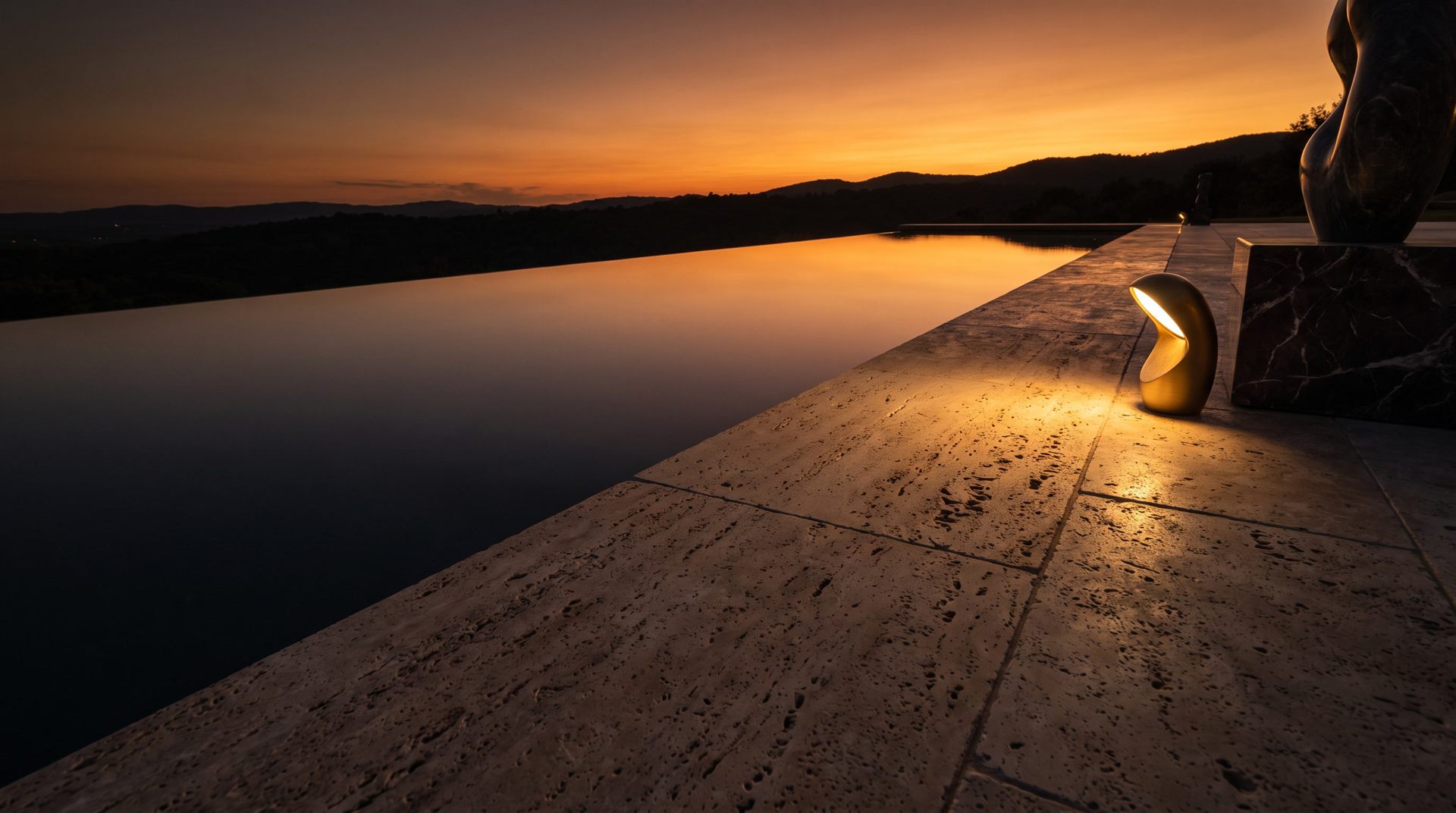 Visione di progetto: piscina a sfioro a filo cielo al tramonto, bordo in travertino romano e lampada d'ottone accesa sulla pietra