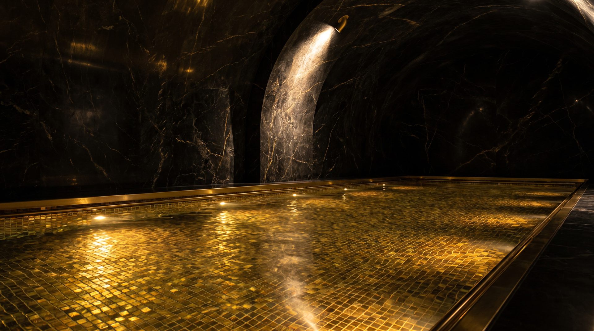 Visione di progetto: vasca interna notturna con fondo di mosaico dorato illuminato, sotto volte di marmo nero venato d'oro